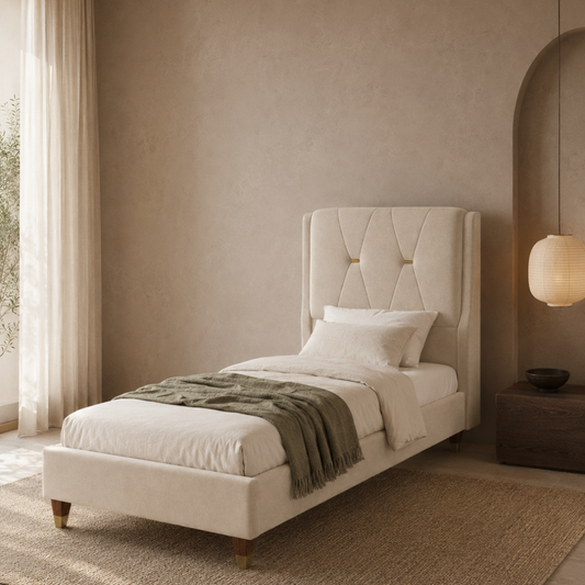 A single bed with a tall tufted upholstered headboard in a warm cream or off-white fabric, styled with white bedding, a textured olive green throw blanket, and wooden legs, set in a minimalist beige bedroom.